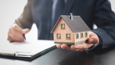 Zbliżenie na dłoń w lewej ręce trzymającej miniaturę domu, w prawej pióro