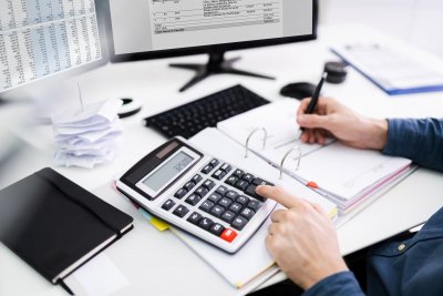 Zbliżenie na dłonie przy biurku przy kalkulatorze, w tle monitory komputerowe