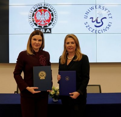 Dwie kobiety stoją w sali konferencyjnej, trzymają dokumenty, w tle logo CBA i Uniwersytetu.