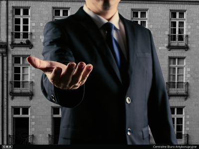 Mężczyzna wyciągający przed siebie dłoń. W tle kamienica.