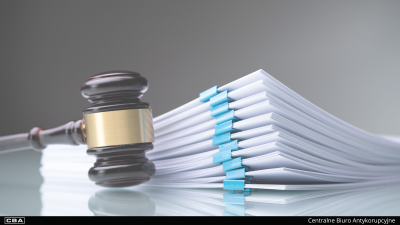 Drewniany młotek sędziowski leżący obok grubego stosu białych dokumentów spiętych klipsami biurowymi. Na dole czarny pasek z logo CBA.