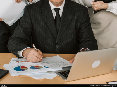 Mężczyzna w garniturze siedzący przy stole, za nim dwie kobiety. Na stole laptop i dokumenty. Na dole czarny pasek z napisem "Centralne Biuro Antykorupcyjne".