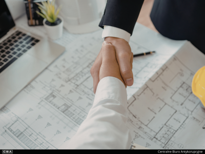 Uścisk dłoni dwóch mężczyzn, odbywający się nad rozłożonymi na biurku planami architektonicznymi. W tle widoczny jest laptop, biały kask ochronny. Na samym dole czarny pasek z logo CBA i napis Centralne Biuro Antykorupcyjne.