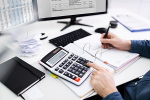A man in a blue shirt performs calculations on a calculator and takes notes, computer monitors in the background