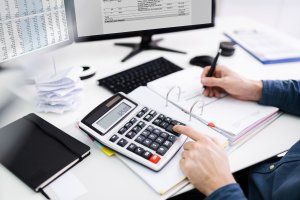 A man in a blue shirt performs calculations on a calculator and takes notes, computer monitors in the background