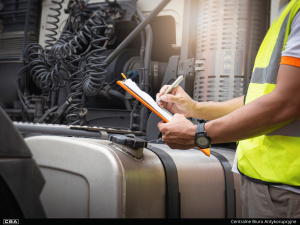 A person in a yellow high-visibility vest stands next to a large truck and writes on a clipboard