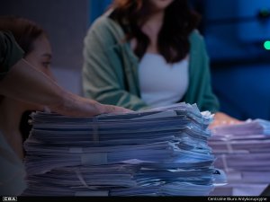 A pile of documents on the desk, a silhouette of a woman in the background.